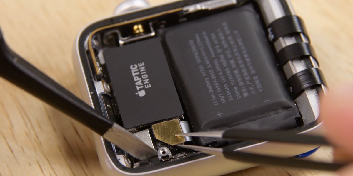 A close-up of a smartwatch being disassembled, with the Taptic Engine and battery visible inside the device