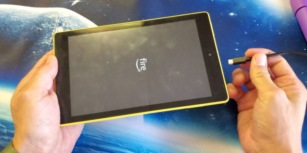 A person holding a yellow Amazon Fire tablet with a charging cable plugged in, displaying the "Fire" logo on the screen