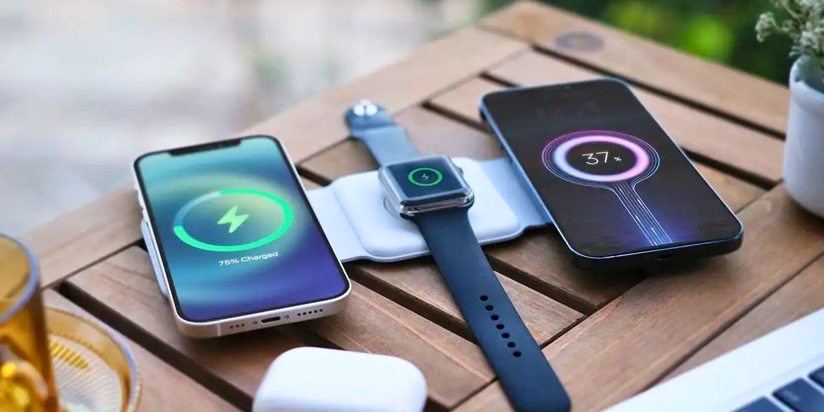 Two smartphones and an Apple Watch are wirelessly charging on a pad on a wooden table.