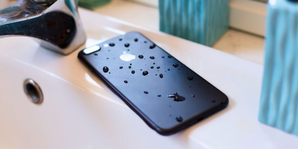 A black smartphone, face down, with water droplets on the back, resting on a bathroom sink.