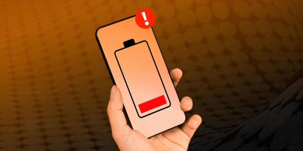 Hand holding a smartphone displaying a low red battery icon and a red exclamation mark warning.