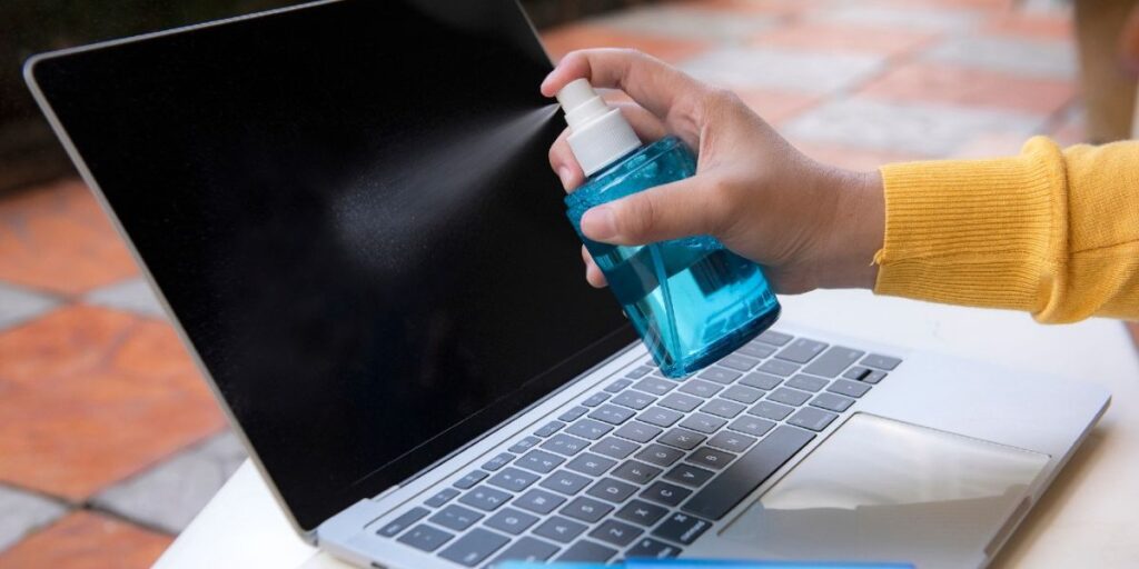 A hand spraying cleaning solution from a blue bottle onto the black screen of an open laptop