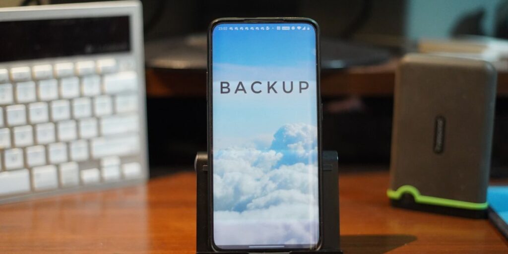 A smartphone on a stand with the word "BACKUP" displayed over a background of white clouds and blue sky