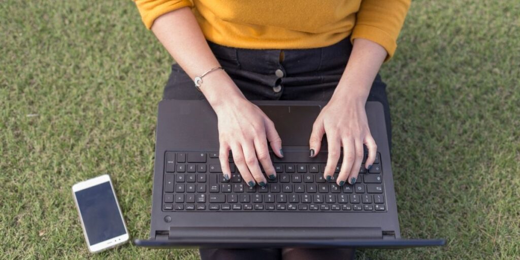 A person in a yellow sweater sits on grass using a black laptop with a white smartphone lying nearby on the lawn